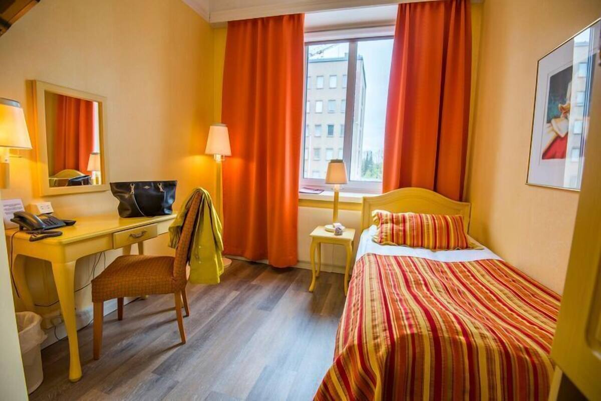 A hotel room with a bed, a desk, a chair, a window with curtains, and a lamp.