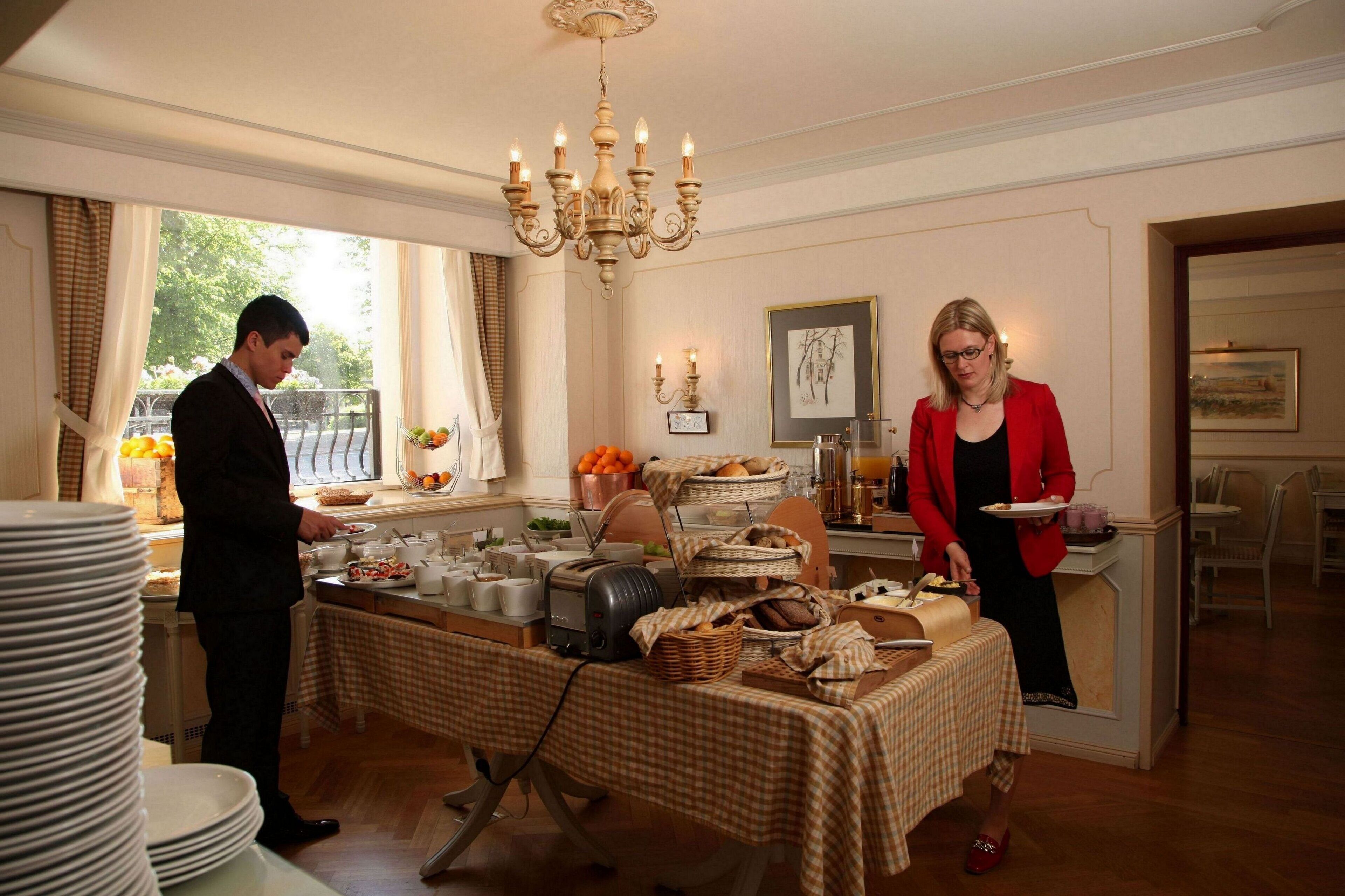 Un buffet de petit-déjeuner est installé sur une table recouverte d’une nappe à carreaux, un lustre illumine l’ensemble, et une fenêtre avec des rideaux complète le décor.