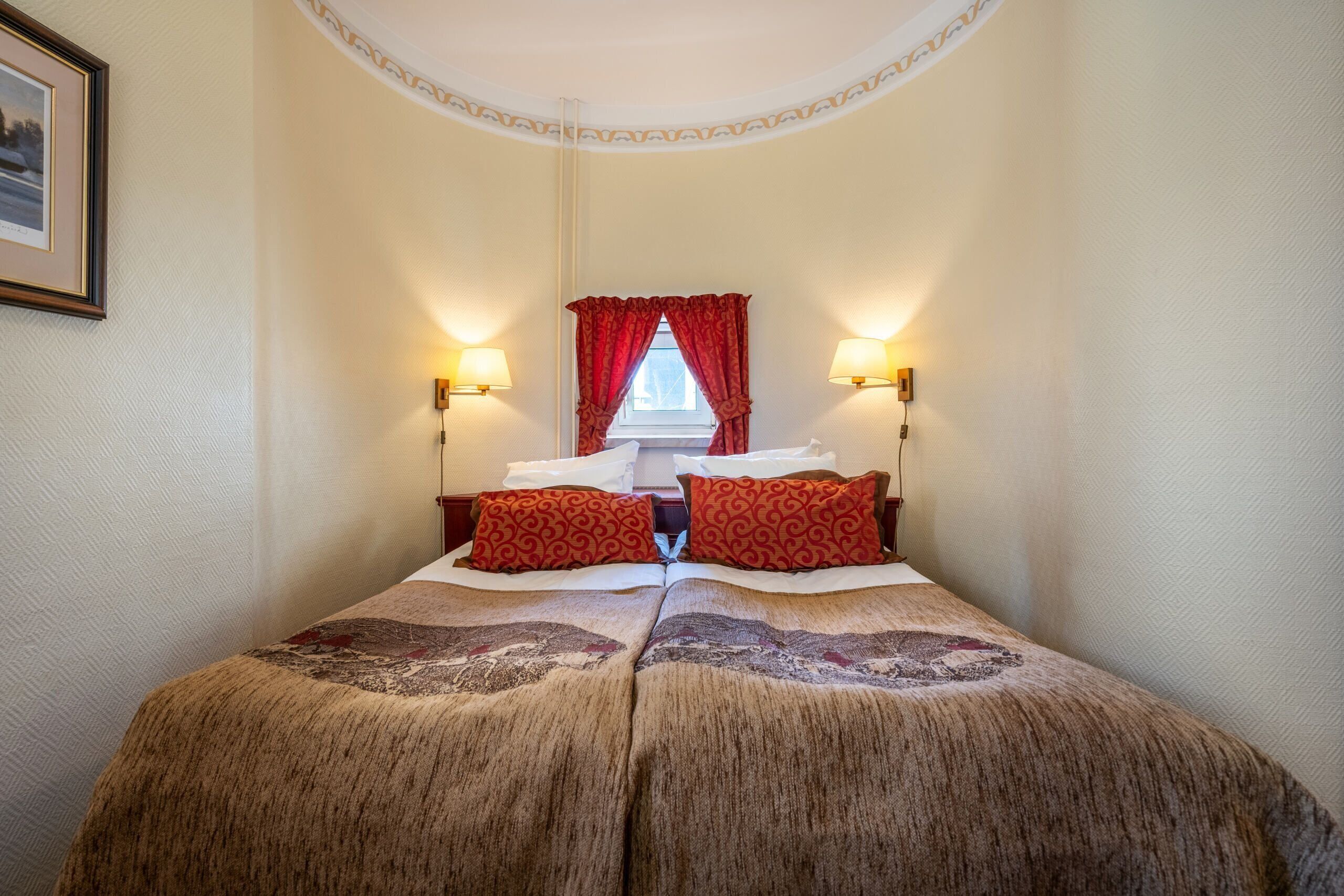 Habitación de hotel con una cama grande, dos almohadas rojas estampadas, dos lámparas de pared, una ventana con cortinas rojas y un cuadro enmarcado en la pared.