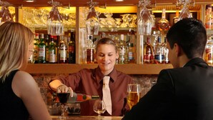 A bar with a bartender preparing drinks, shelves stocked with bottles, and hanging glassware. - Hotel Astor (Vaasa)