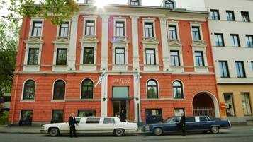 Un edificio rojo con el letrero "ASTOR" en la fachada. Hay dos coches estacionados adelante y una persona parada cerca de la entrada.