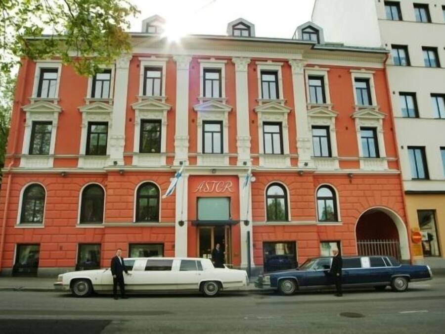 Un edificio rojo con el letrero "ASTOR" en la fachada. Hay dos coches estacionados adelante y una persona parada cerca de la entrada.