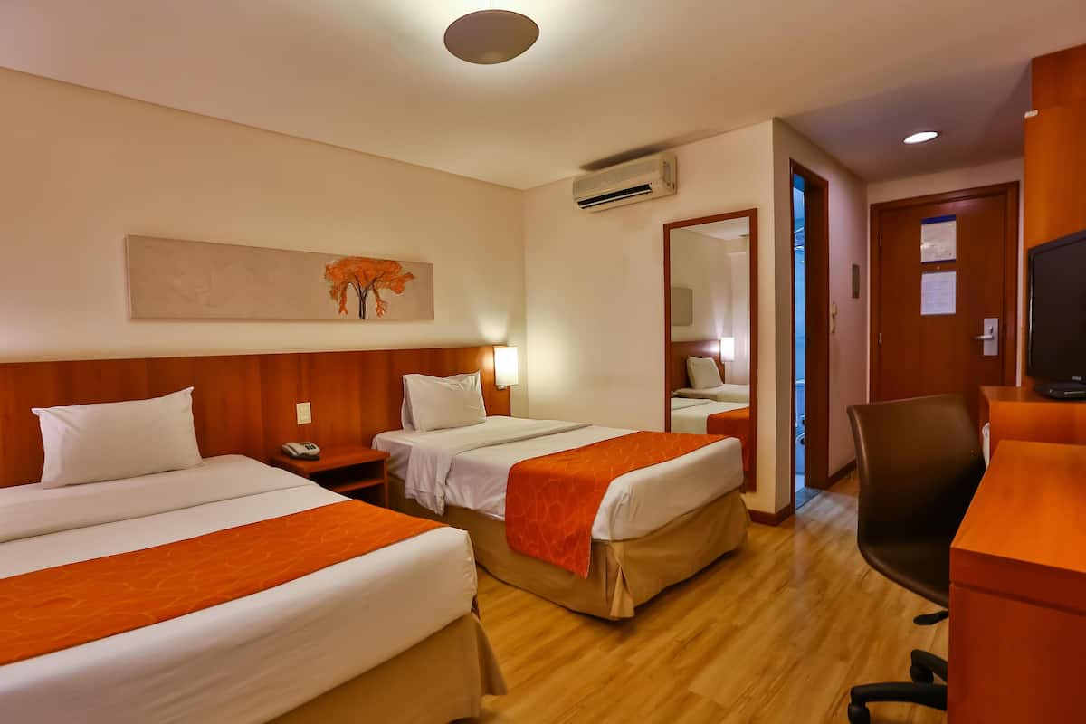 A hotel room with two beds, a desk, a chair, an air conditioner, and a painting on the wall.