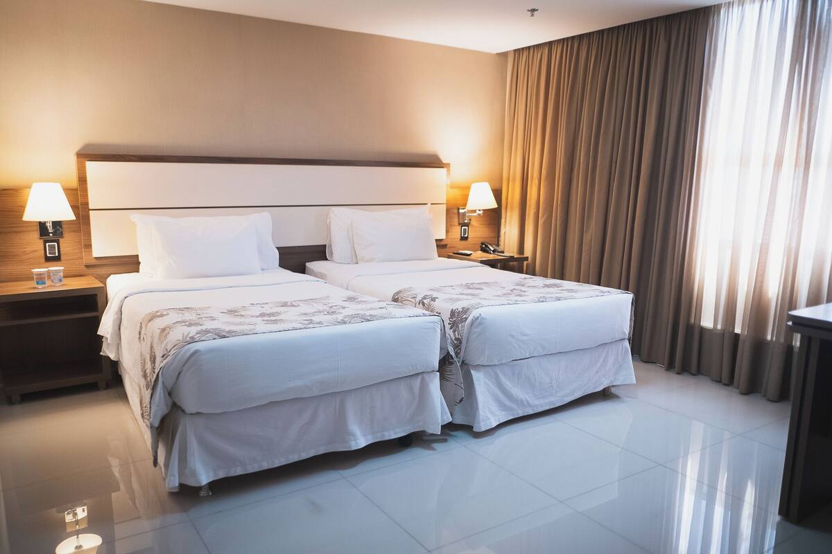 A hotel room with two beds, a large headboard, bedside lamps, a nightstand, and a view of the outside through curtains.
