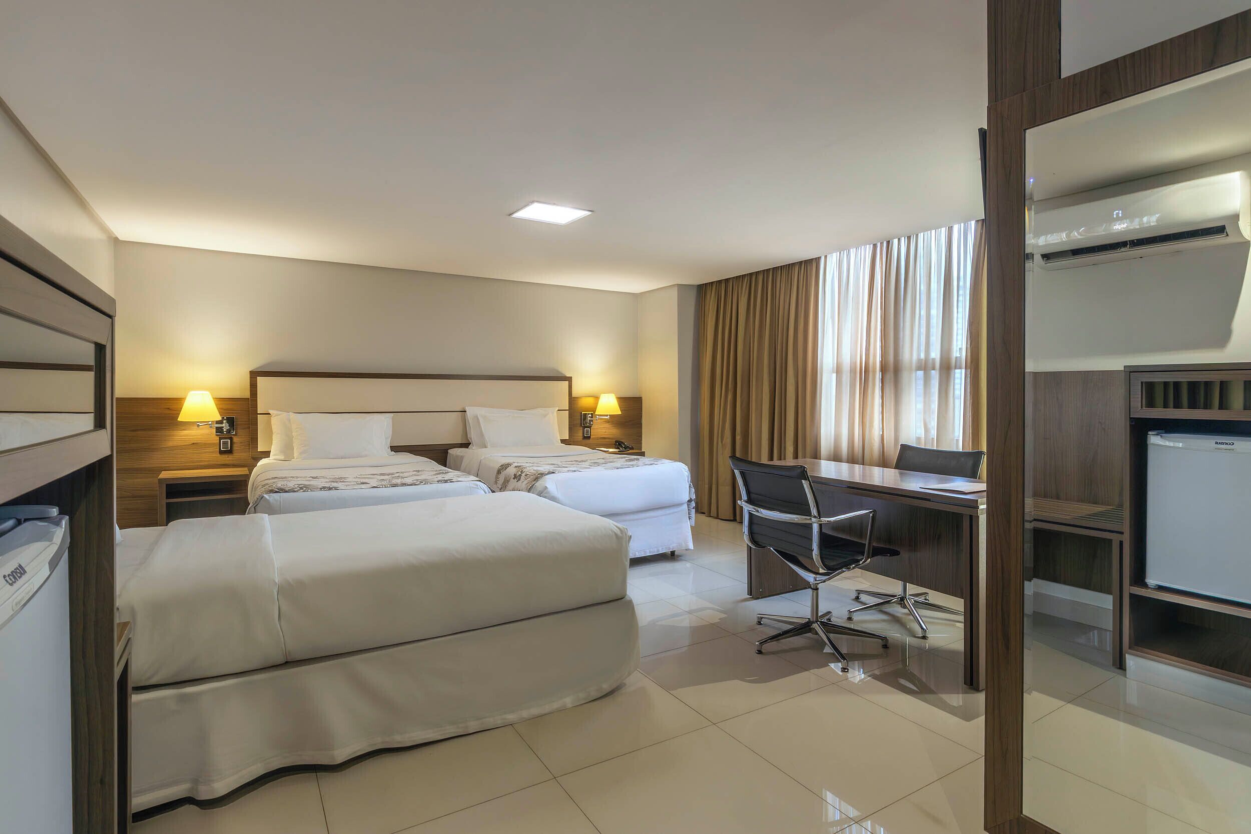 A hotel room with two beds, a desk, a chair, an air conditioner, and a minibar.