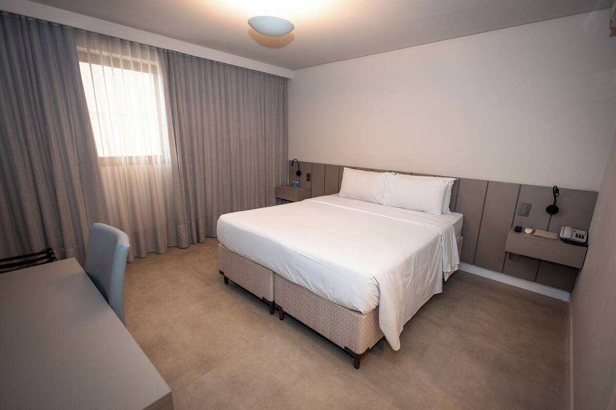 A hotel room with a large bed, a desk, a chair, a nightstand, and a window with curtains.