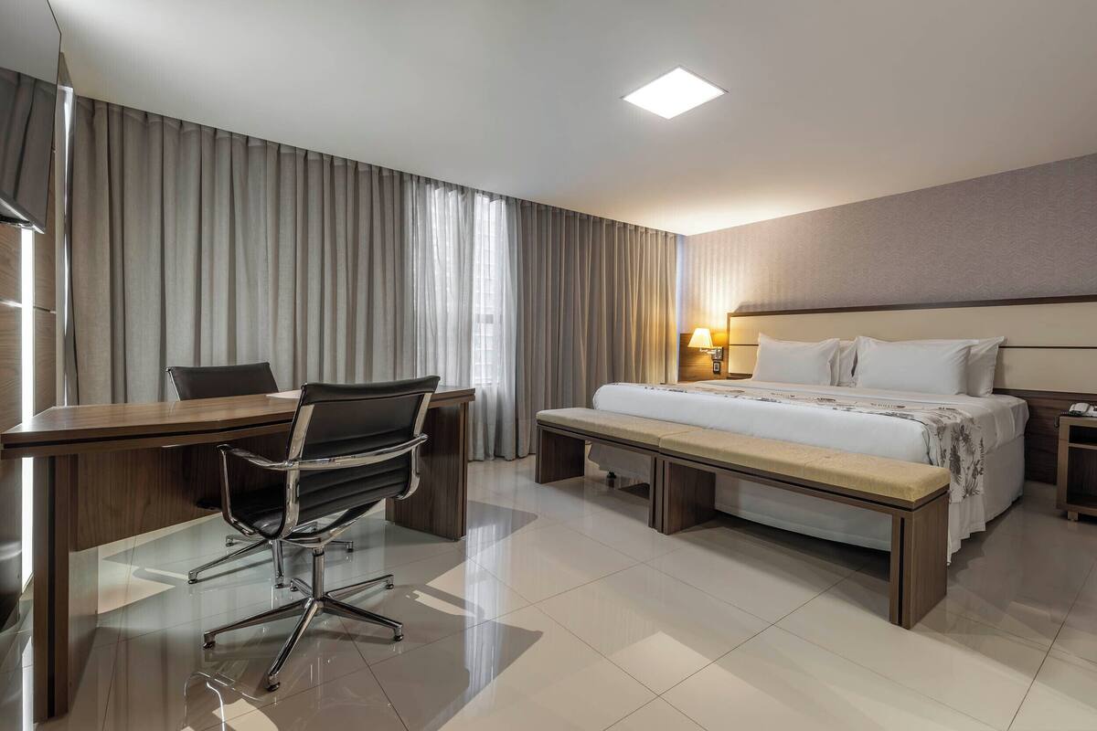 A hotel room with a bed, a desk with a chair, a bench, a bedside table with a lamp, and a large window with curtains.