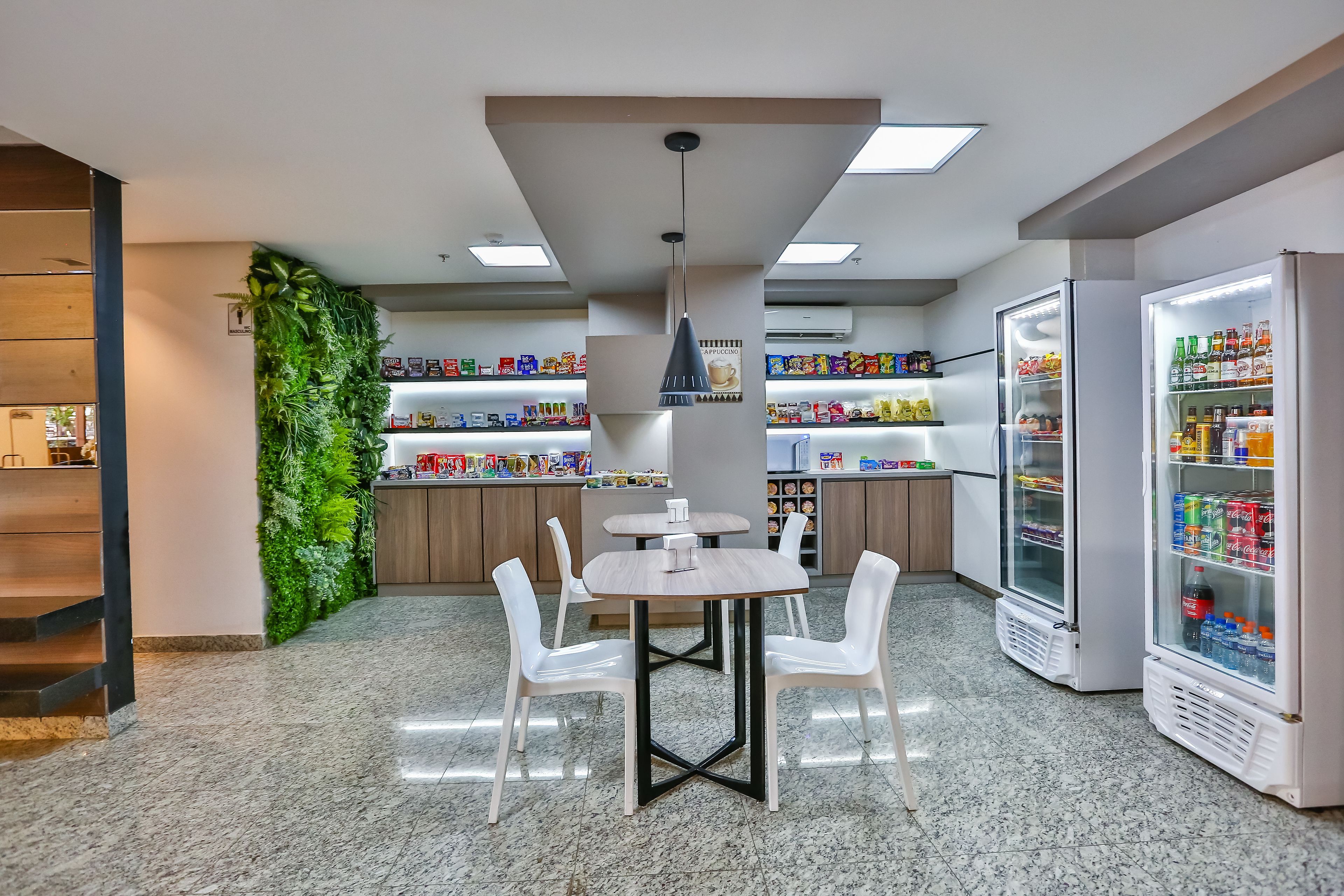 Um café moderno com mesa de madeira e cadeiras brancas, rodeado por prateleiras repletas de lanches e bebidas.