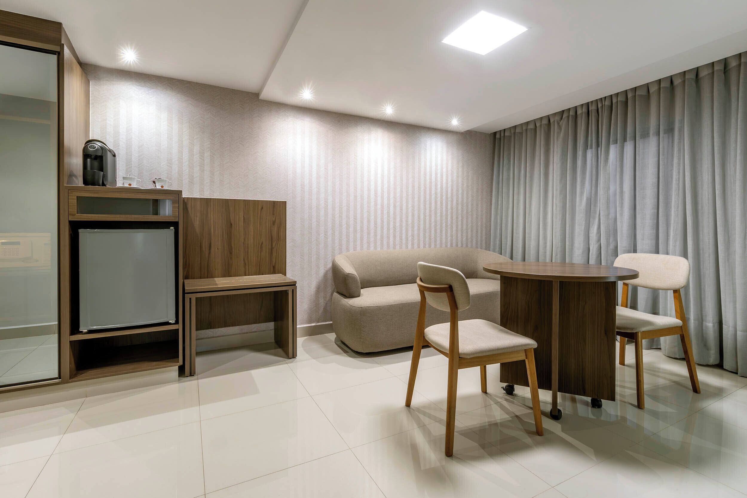 A modern hotel room with a sofa, two chairs, a small table, a coffee machine, and a mini-fridge.