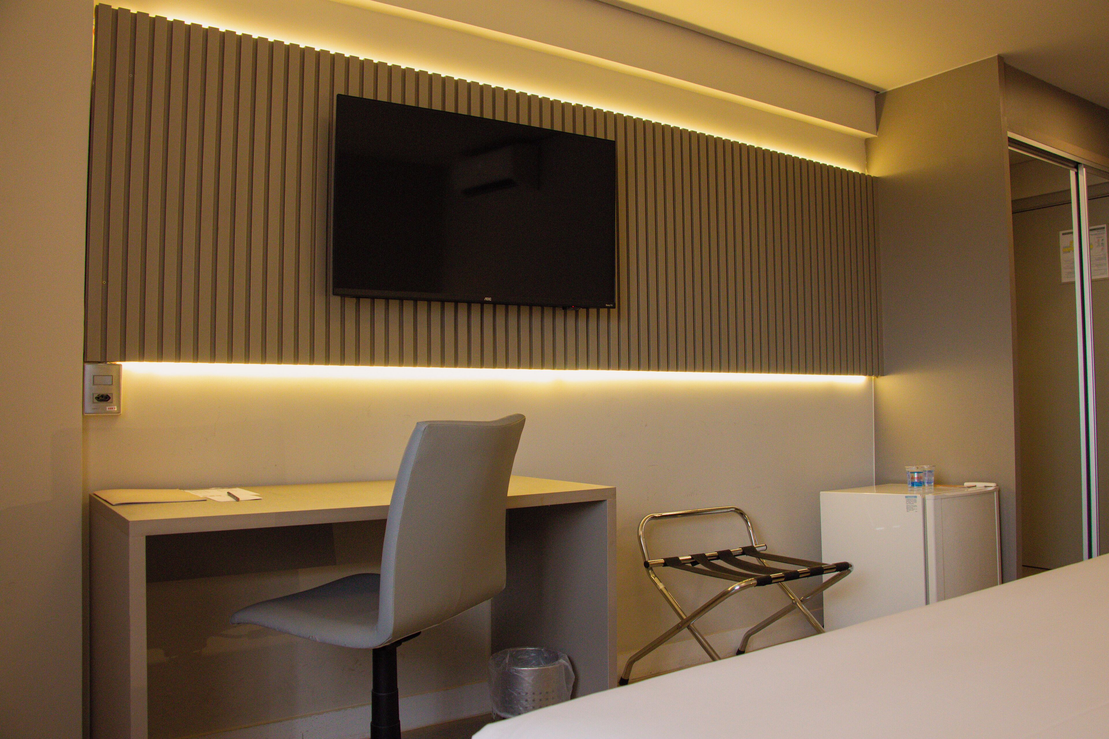A hotel room with a bed, a desk, a chair, a television mounted on the wall, and a small refrigerator.