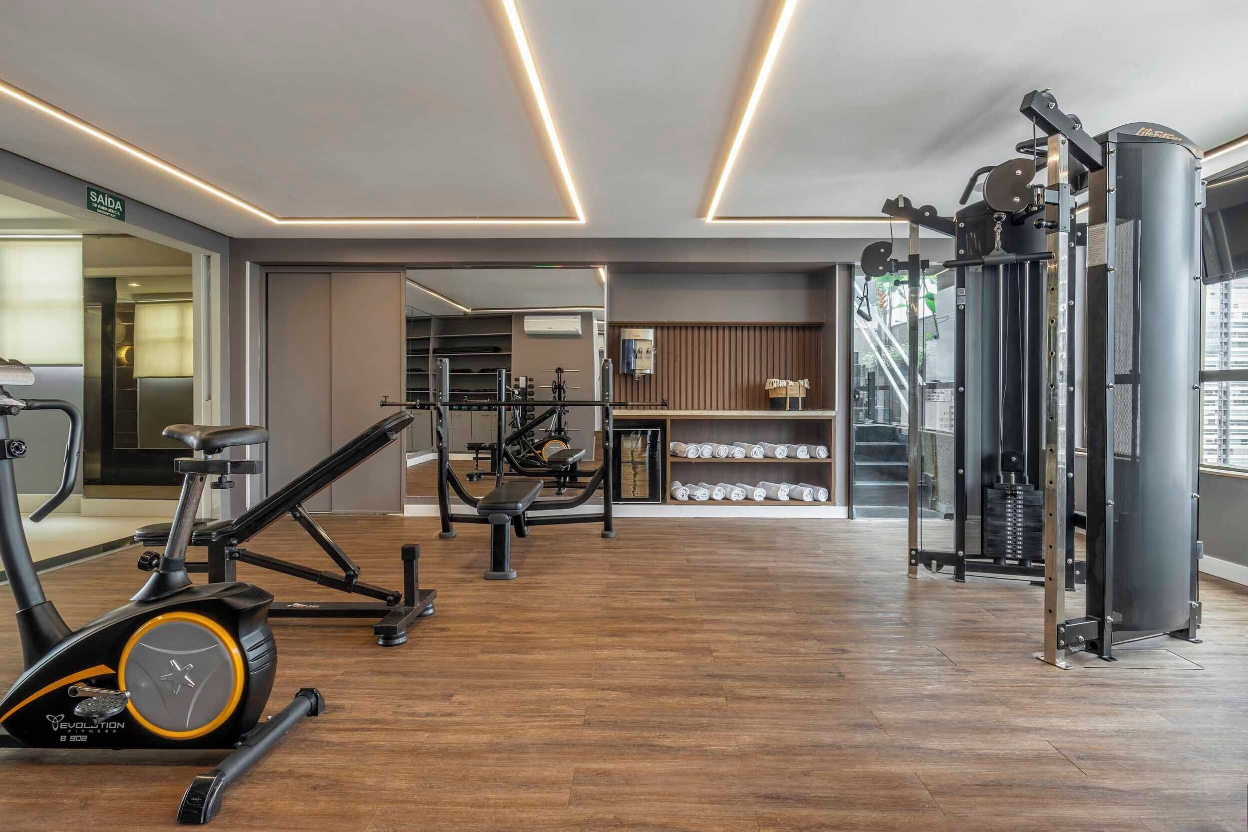 Uma academia com equipamentos de ginástica, esteira e um suporte de halteres na parede.