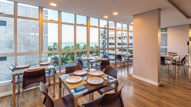 A modern dining area with large windows, wooden flooring, and a view of the cityscape. - Hotel Costamar Ponta Verde (Maceió)