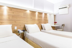 A hotel room with two beds, a bedside table with a phone, a small table with a vase of flowers, and an air conditioning unit on the wall.