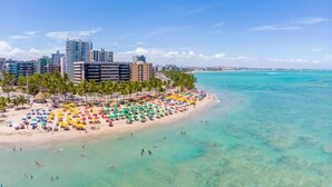 A beach with clear turquoise waters, a row of colorful umbrellas, and a cityscape in the background. - Hotel Costamar Ponta Verde (Maceió)