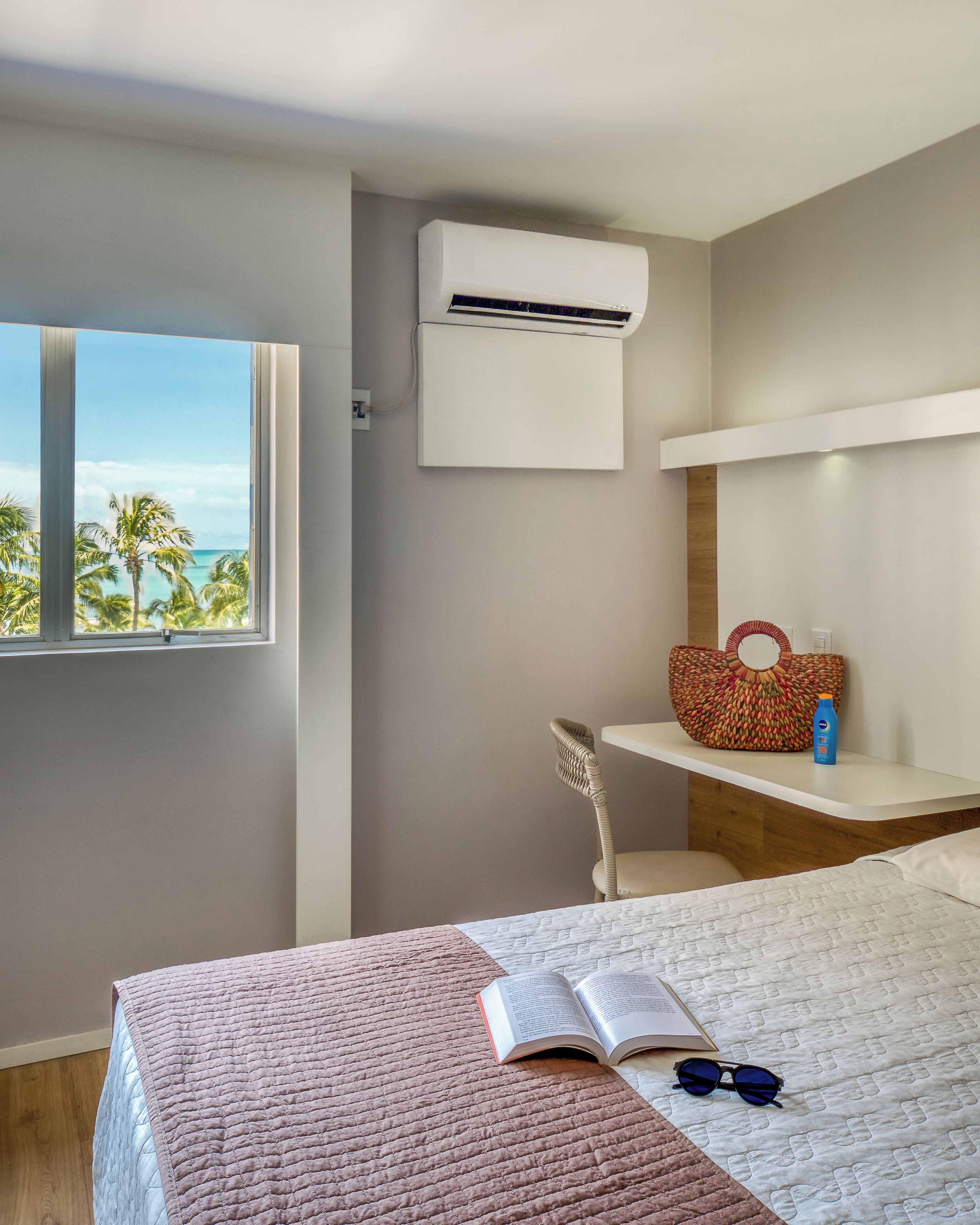 Un dormitorio con cama, ventana con vista a palmeras y al océano, aire acondicionado de pared, una silla de mimbre y una estantería con un bolso y una botella.