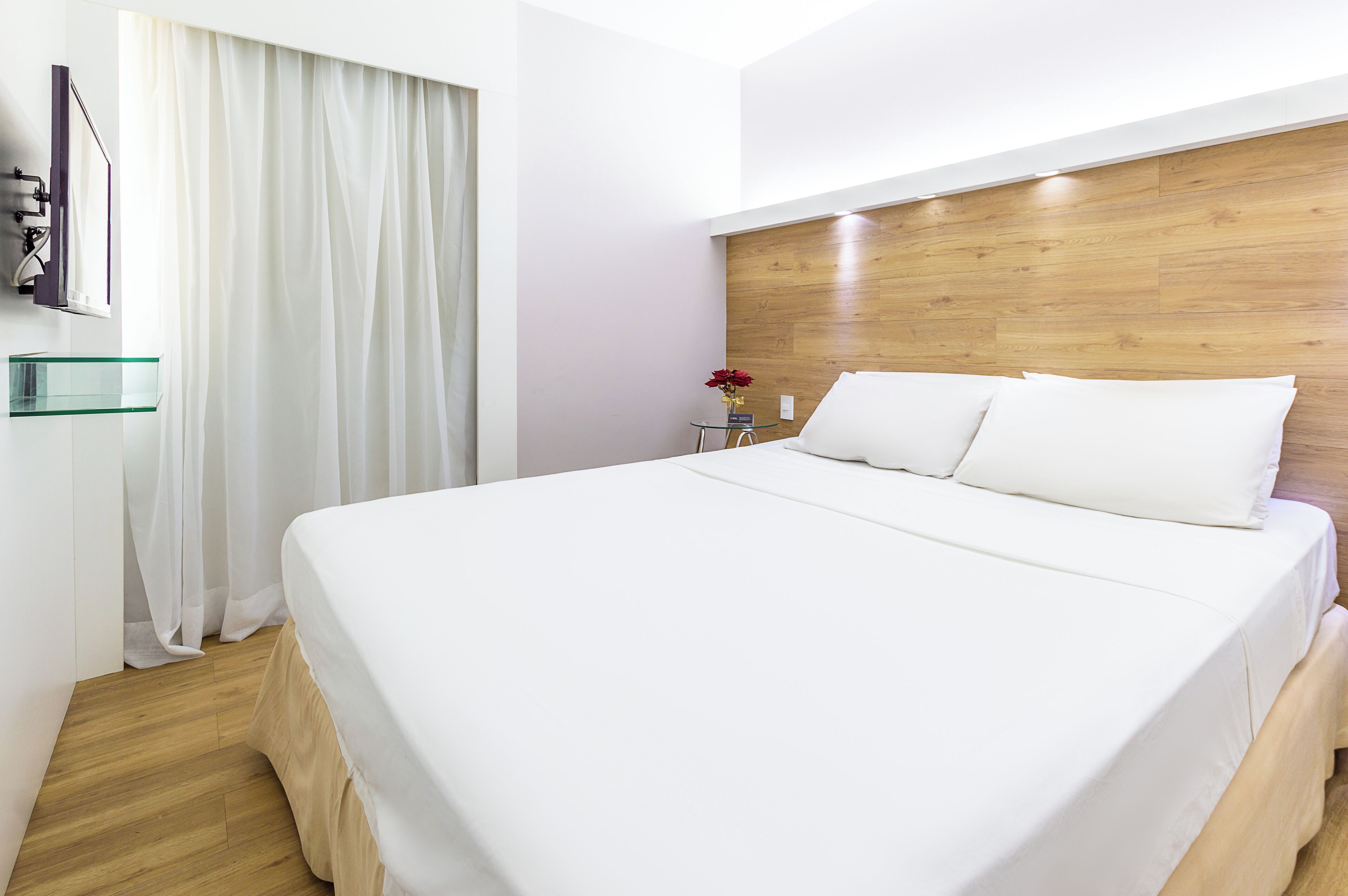 A modern hotel room with a large bed, wooden headboard, and a glass bedside table with a vase of flowers.