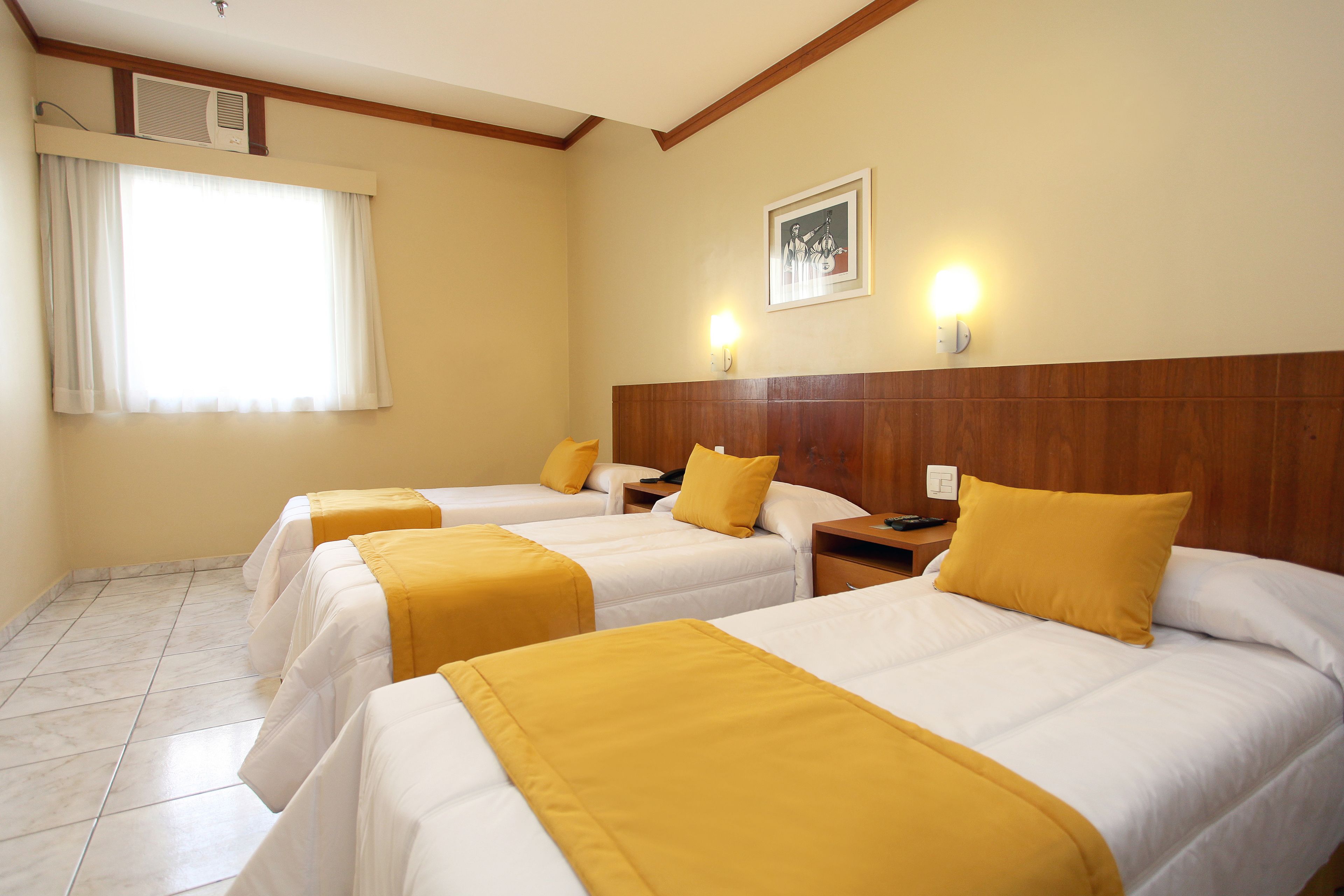 A hotel room with three beds, each with a yellow bedspread and white bedding. There is a wooden headboard, a framed picture on the wall, and a window with curtains.
