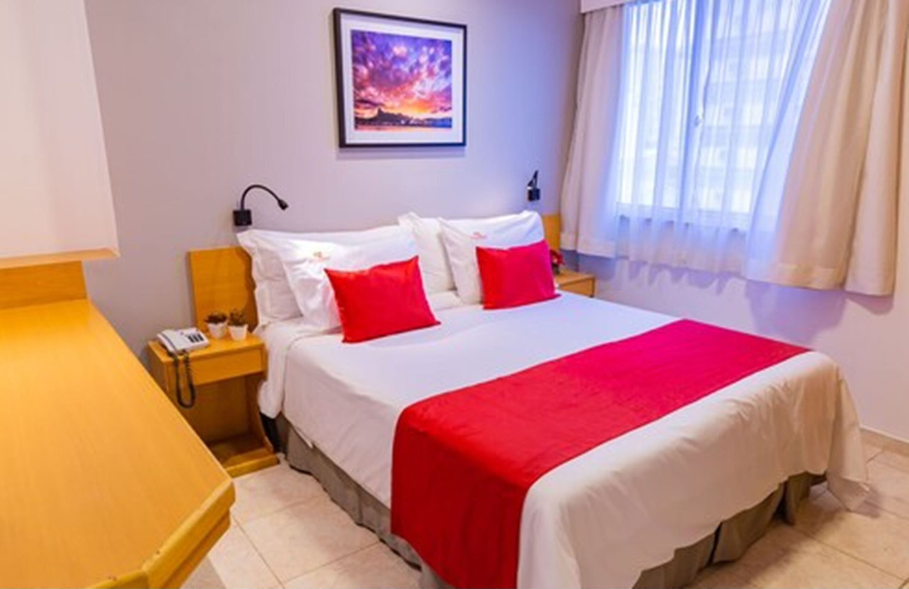 A hotel room with a bed, a nightstand, a lamp, a painting on the wall, and a window with curtains.