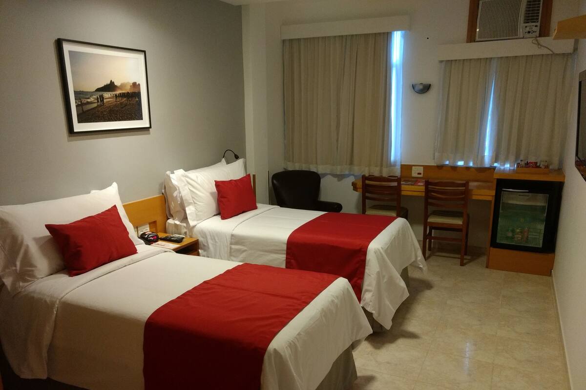 A hotel room with two beds, a desk, a chair, a refrigerator, and a window with curtains.