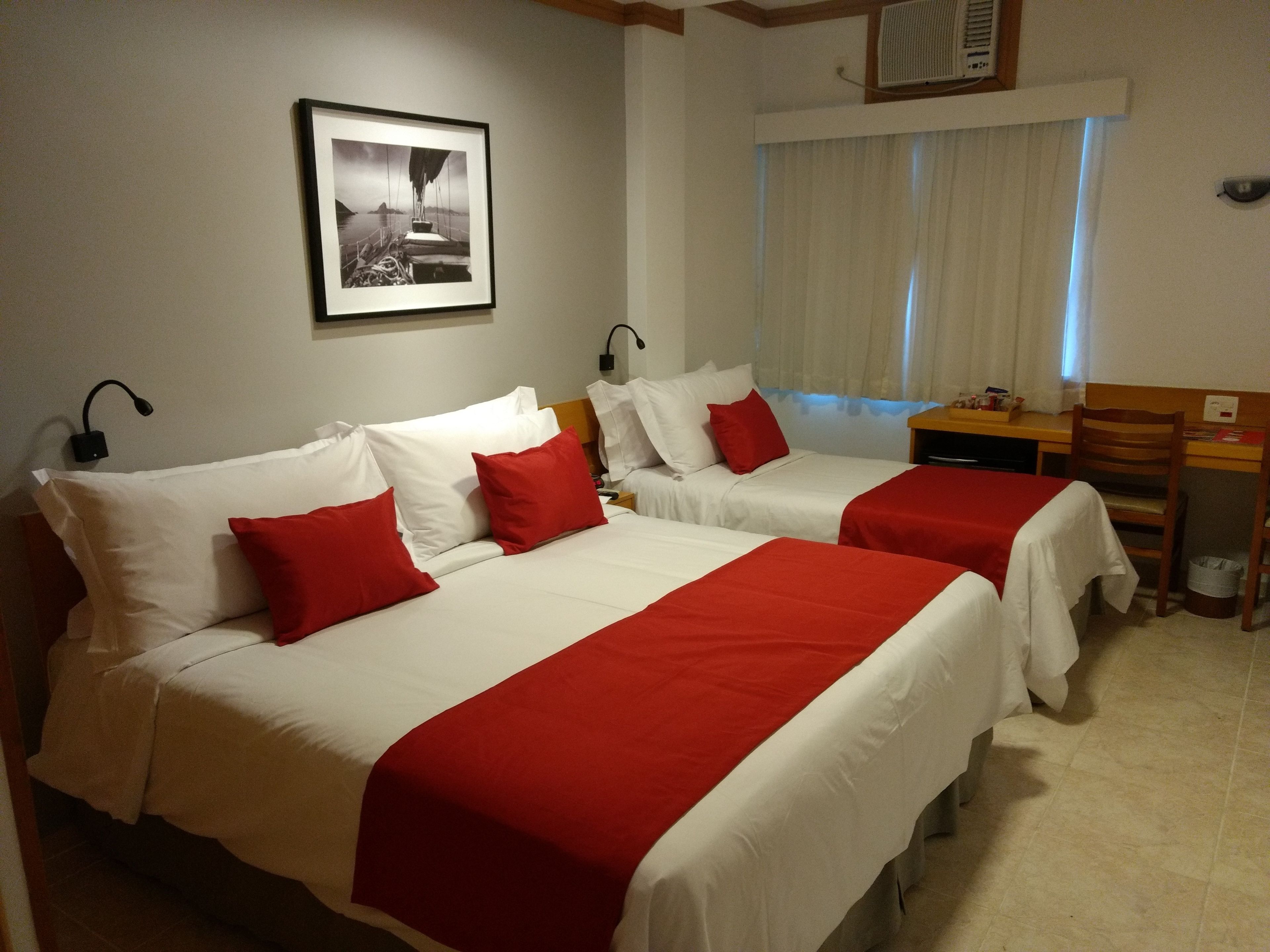 A hotel room with two beds, each with red and white bedding, a desk, and a chair.