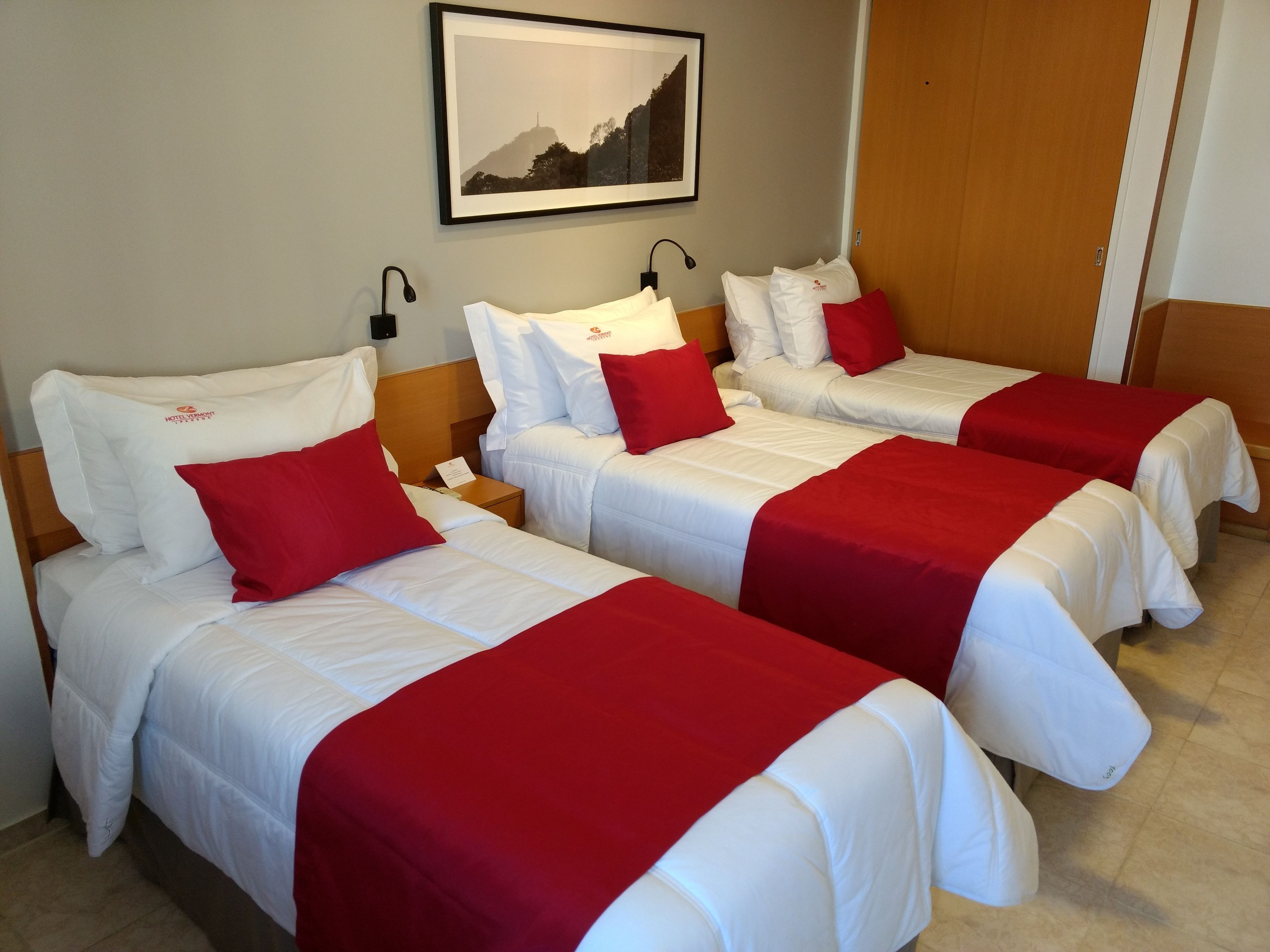 Una habitación de hotel con tres camas, cada una con ropa blanca y detalles rojos. Hay un cuadro enmarcado en la pared sobre las camas.