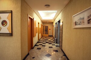 A hotel hallway with a checkered floor, framed artwork on the walls, and a door with a geometric design.
