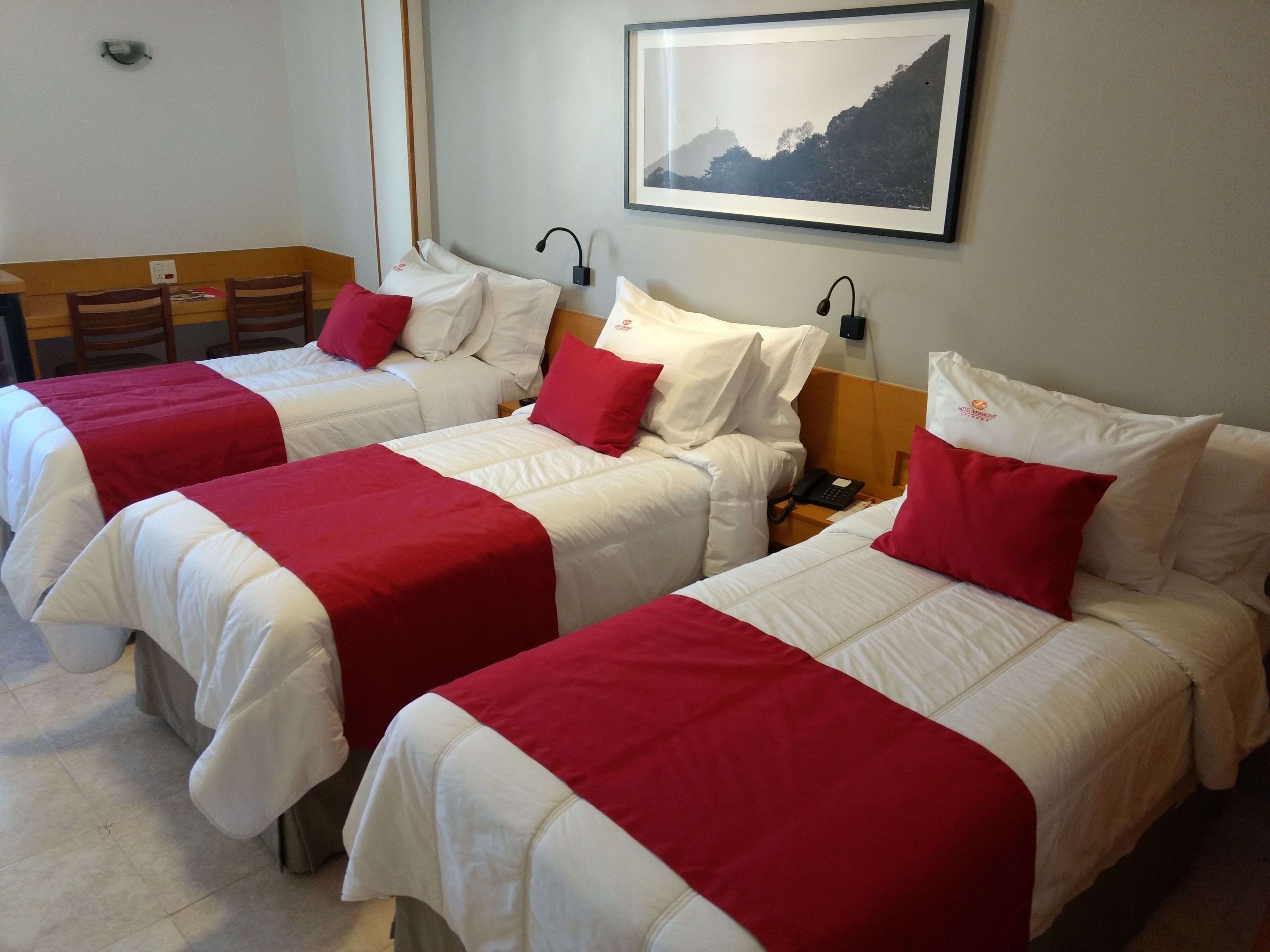 Ein Hotelzimmer mit drei Betten, jedes mit weißer Bettwäsche und roten Akzenten. An der Wand hängt ein gerahmtes Bild, ein Nachttisch mit Lampe und ein Telefon auf dem Nachttisch.