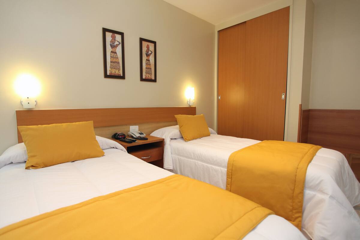 A hotel room with two beds, each with a yellow bedspread and white linens. There are two wall-mounted light fixtures, a wooden headboard, and a wooden door.