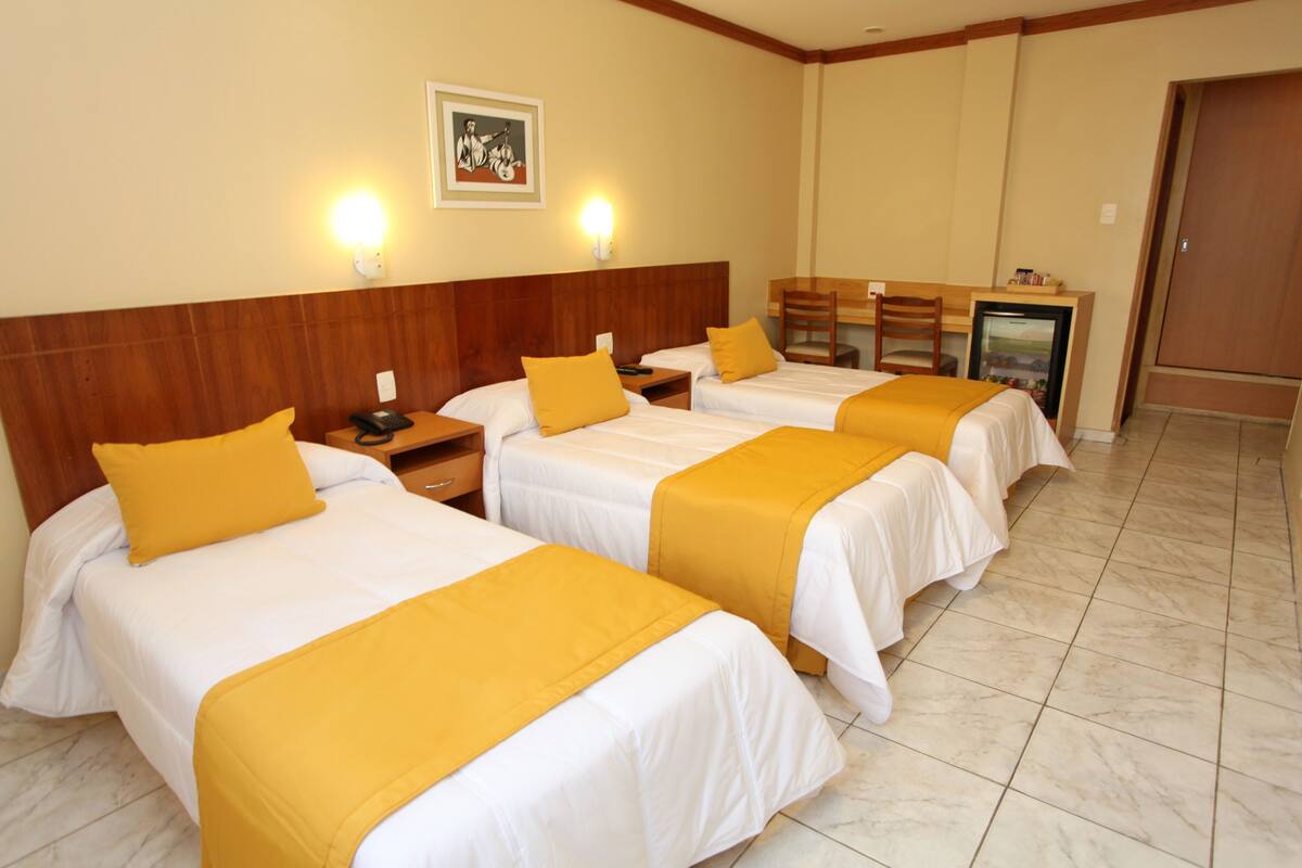 A hotel room with three beds, each with a white bedspread and a yellow accent pillow. There is a wooden headboard, a small desk with a chair, and a refrigerator.