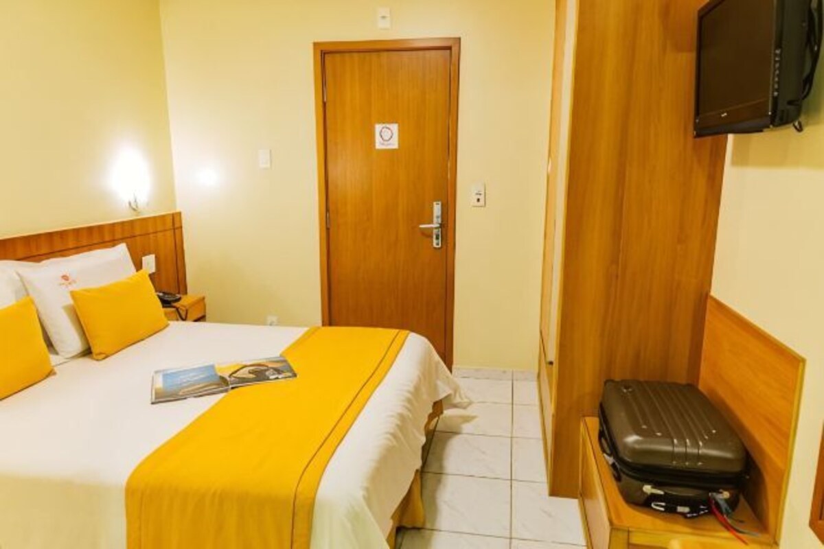 A hotel room with a bed, a wooden headboard, a television mounted on the wall, and a suitcase on a wooden bench.