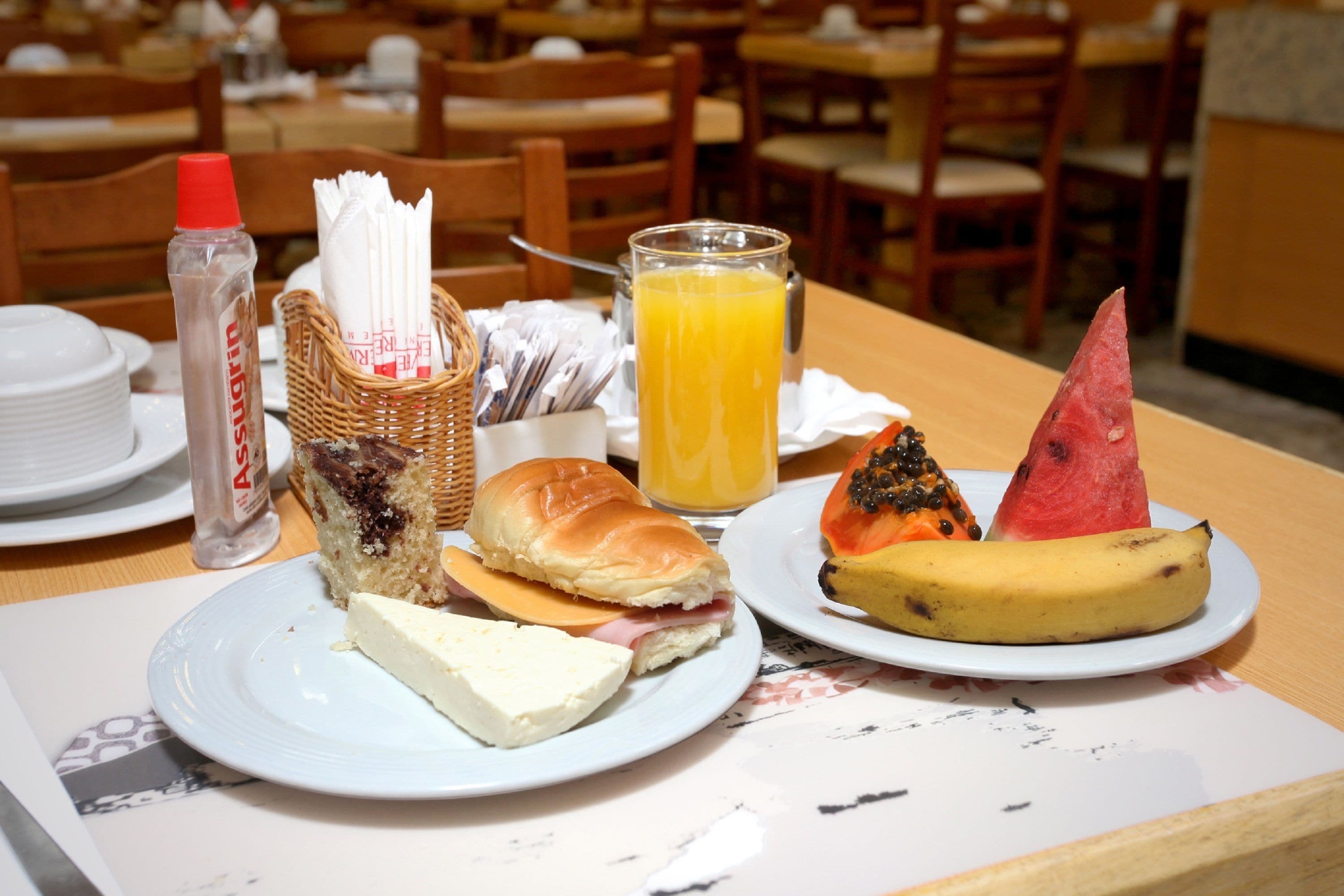 A breakfast setting with a plate of food, a bottle of syrup, a glass of orange juice, and a basket of utensils.