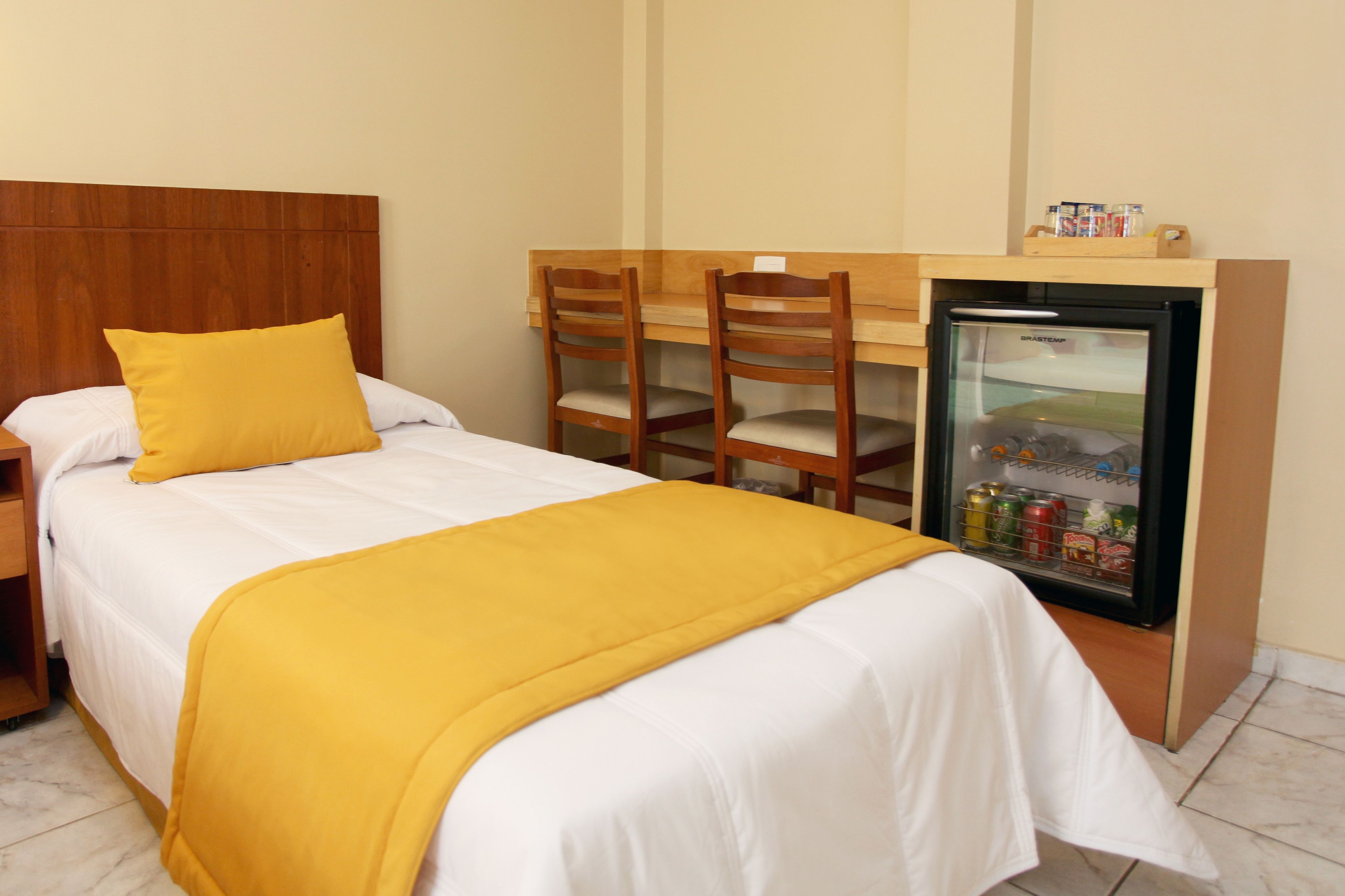 Una habitación de hotel con una cama, un escritorio, dos sillas, un minibar y un cabecero de madera.