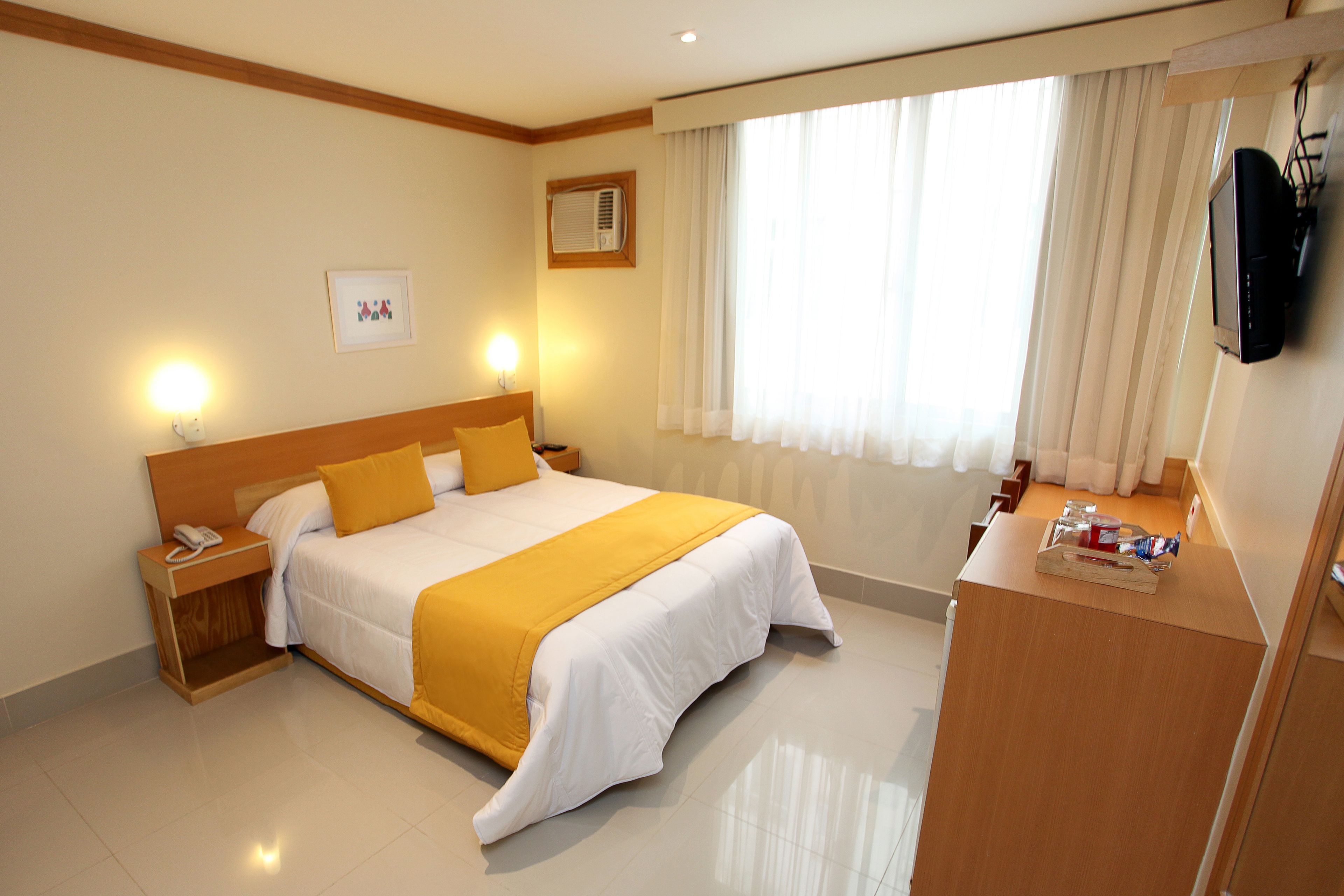 Quarto de hotel com cama, mesa de cabeceira, luminária e televisão na parede.