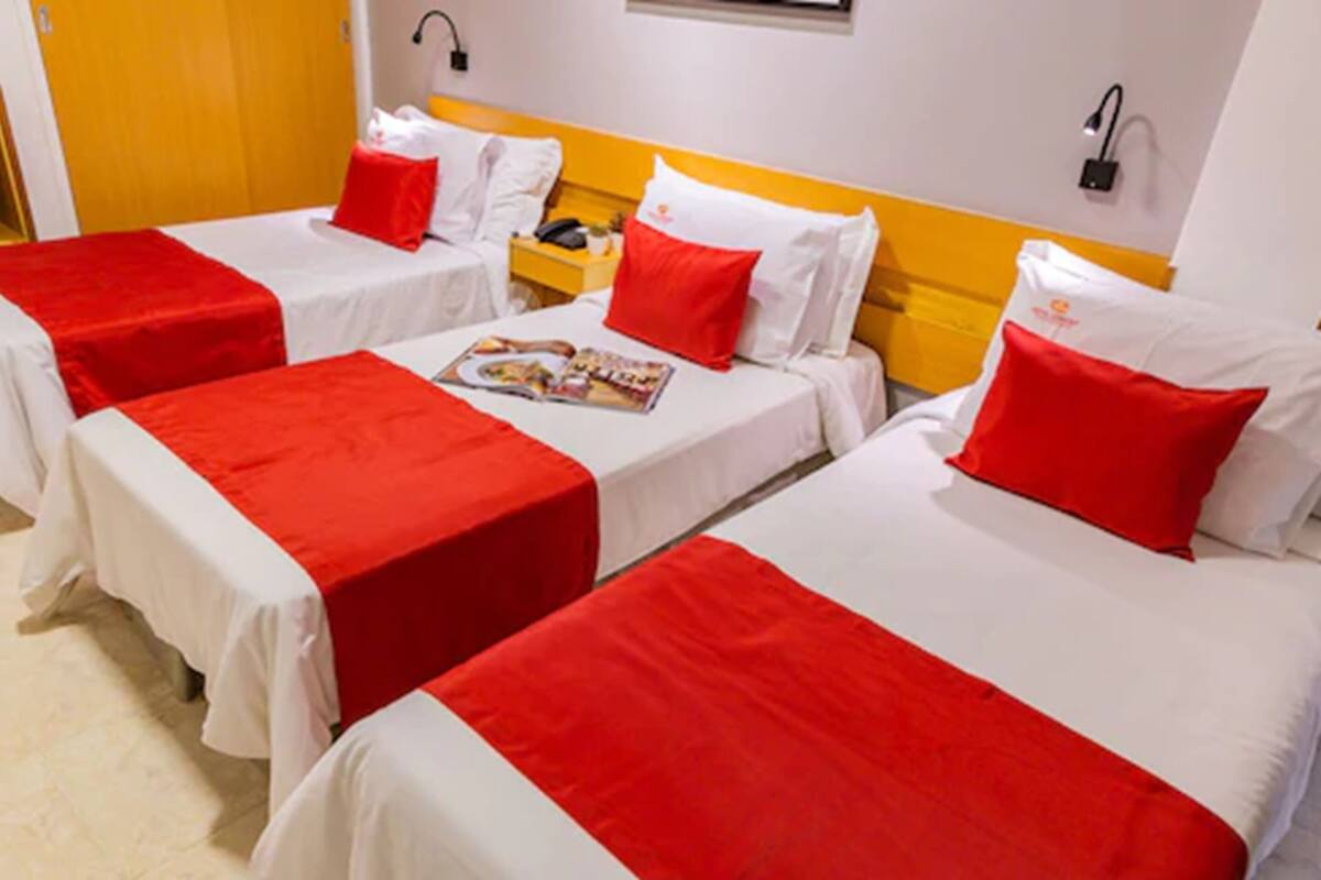 A hotel room with three single beds, each with white bedding and red accents. There is a nightstand with a lamp and a telephone on one side.