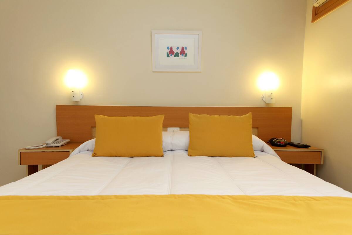 A hotel room with a bed, two bedside tables, a nightstand, a framed picture on the wall, and two wall-mounted lamps.