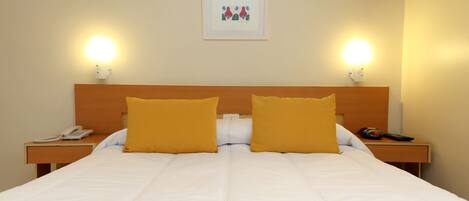 A hotel room with a bed, two bedside tables, a nightstand, a framed picture on the wall, and two wall-mounted lamps.
