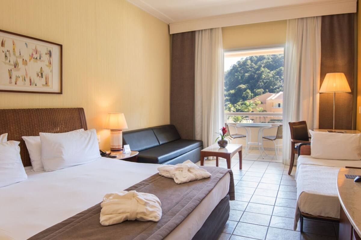 A hotel room with a large bed, a sofa, a desk, and a balcony with a view of the trees.