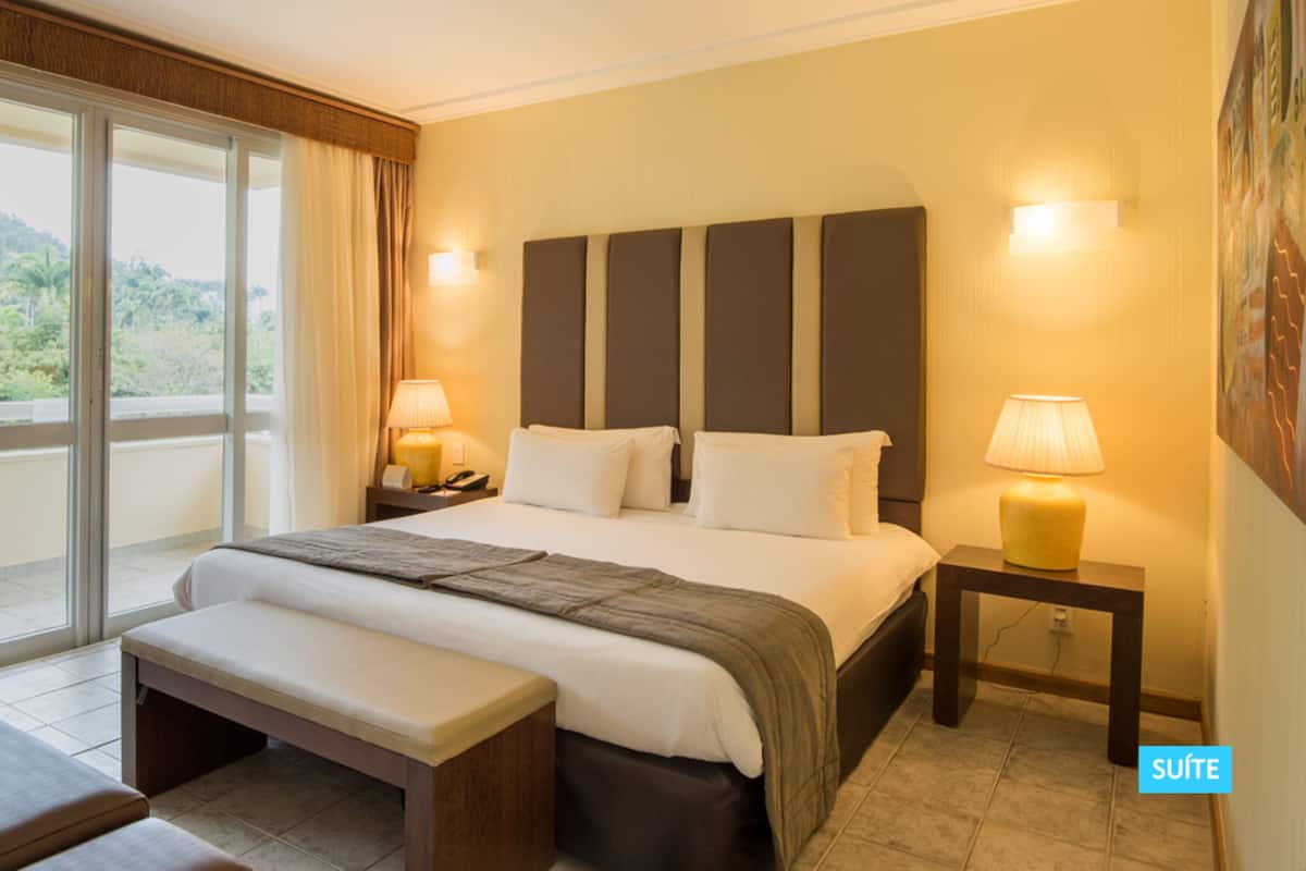 A hotel room with a large bed, a bench, a desk, and a view of greenery through the window.