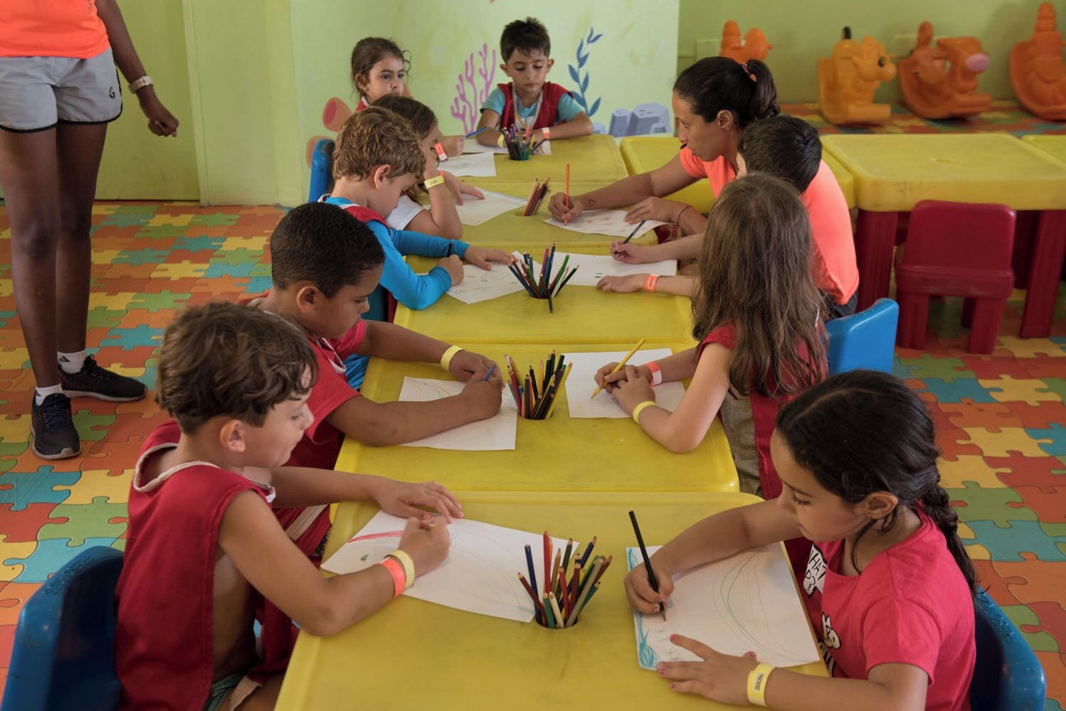 Crianças participando de atividades de desenho em uma mesa com lápis coloridos, em um quarto com um espaço de recreação ao fundo.