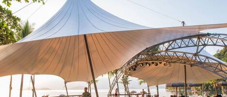 Un espacio frente a la playa con una gran estructura tipo toldo y un escenario para eventos.