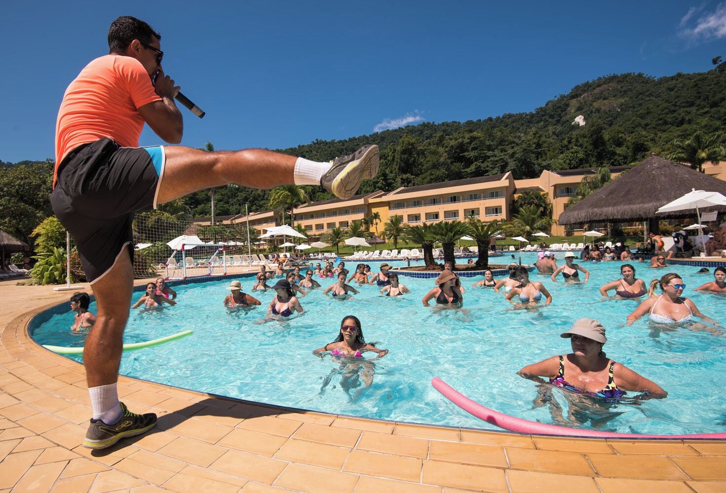 A swimming pool area with people in the water, a person standing on a board, and a building in the background.