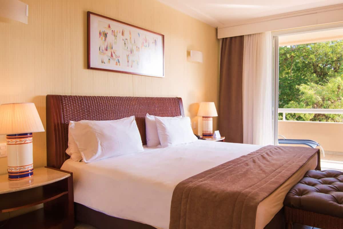 A hotel room with a large bed, two bedside lamps, a television on a stand, and a balcony with a view of greenery.