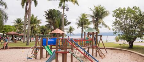 A playground with slides and climbing structures, surrounded by palm trees and a beach.