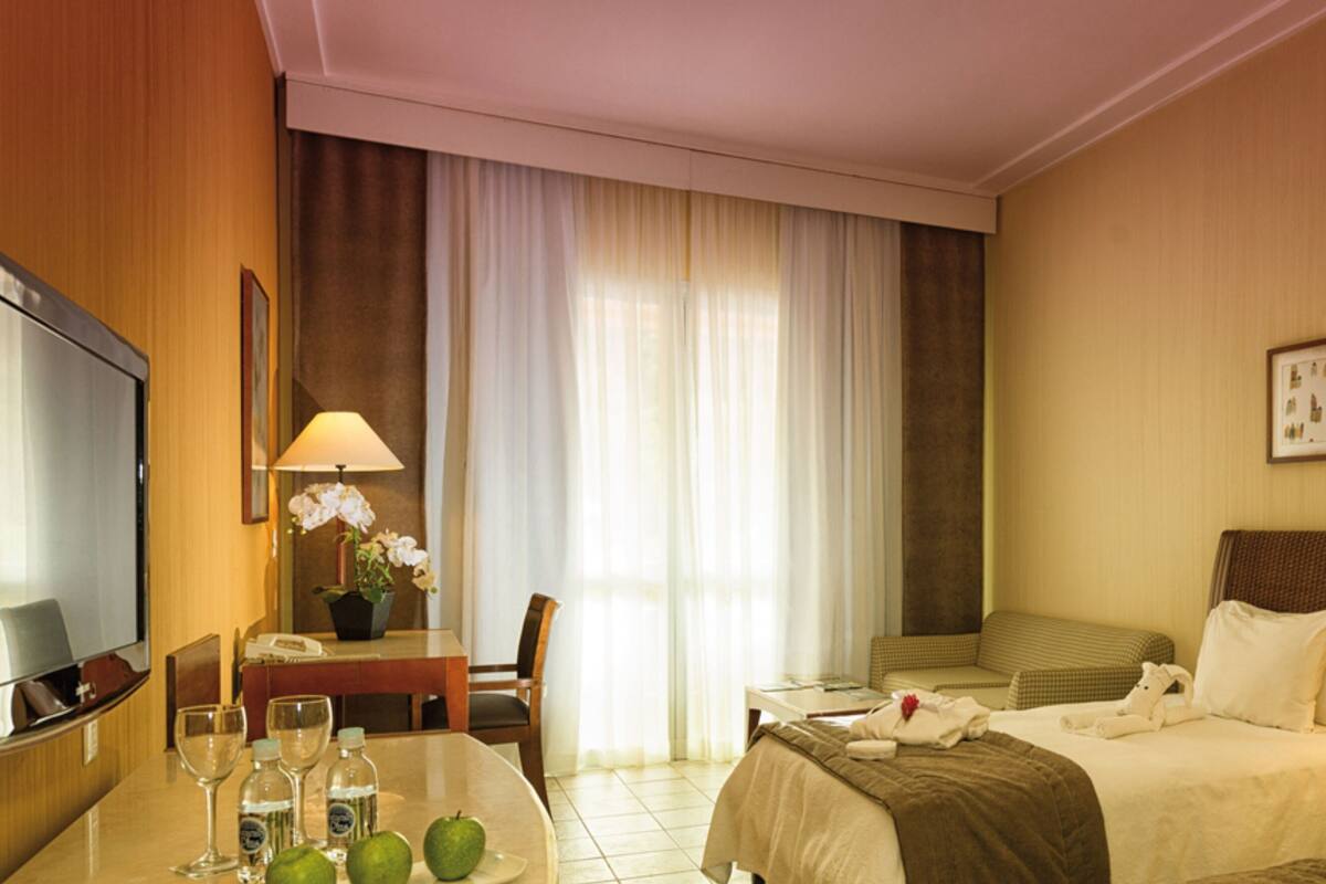 A hotel room with a TV, two beds, a desk with a lamp, a chair, and a window with curtains.