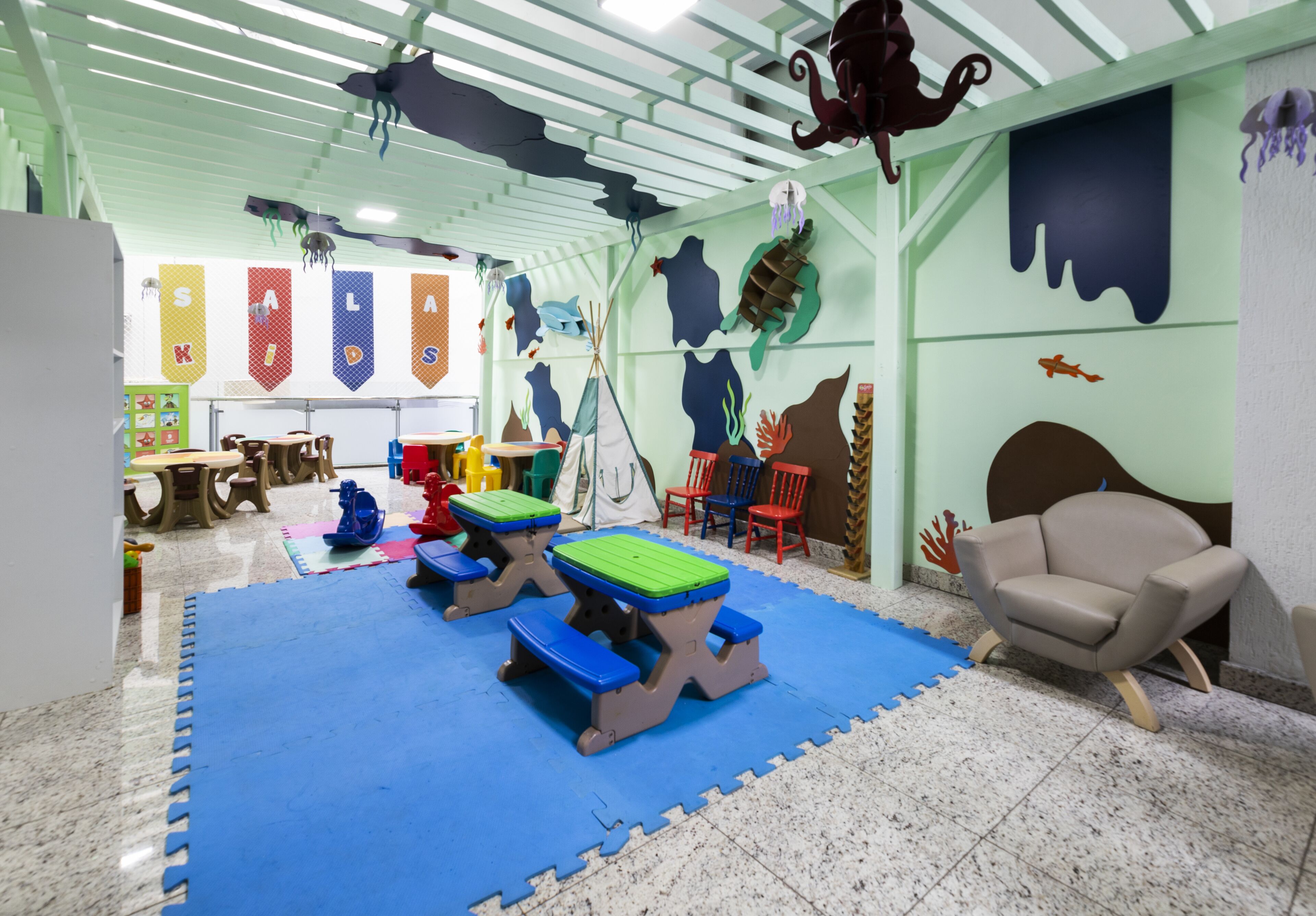 Zona de juegos infantiles con colchonetas azules, mesas de picnic y una silla color beige.
