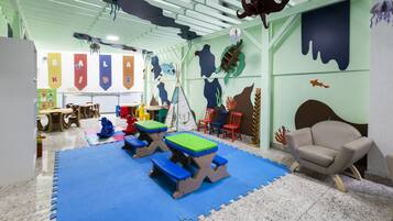 Zona de juegos infantiles con colchonetas azules, mesas de picnic y una silla color beige.