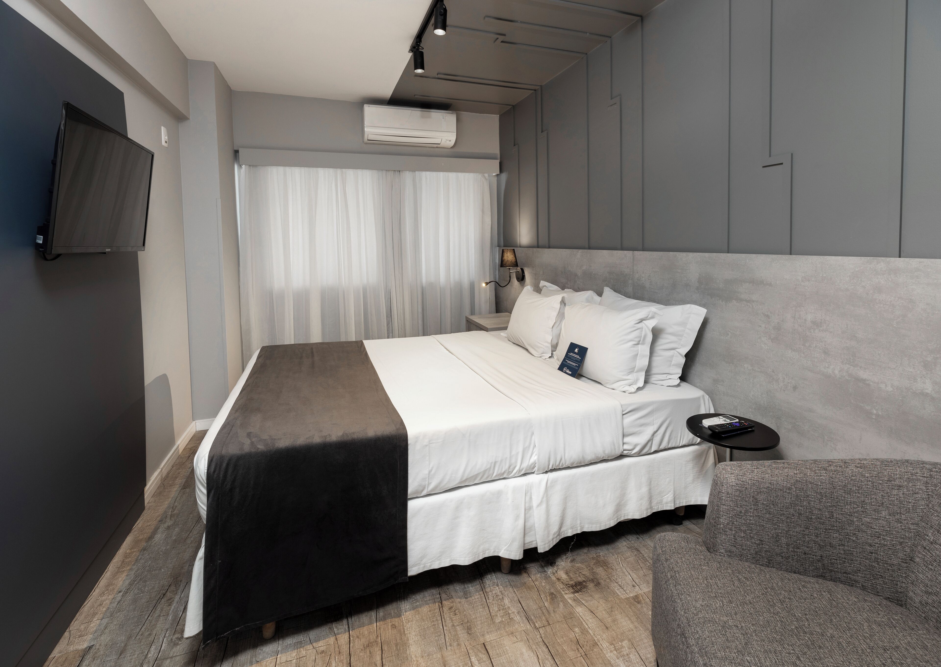 A modern hotel room with a large bed, a flat-screen TV mounted on the wall, a bedside table with a remote control, and a grey upholstered armchair.