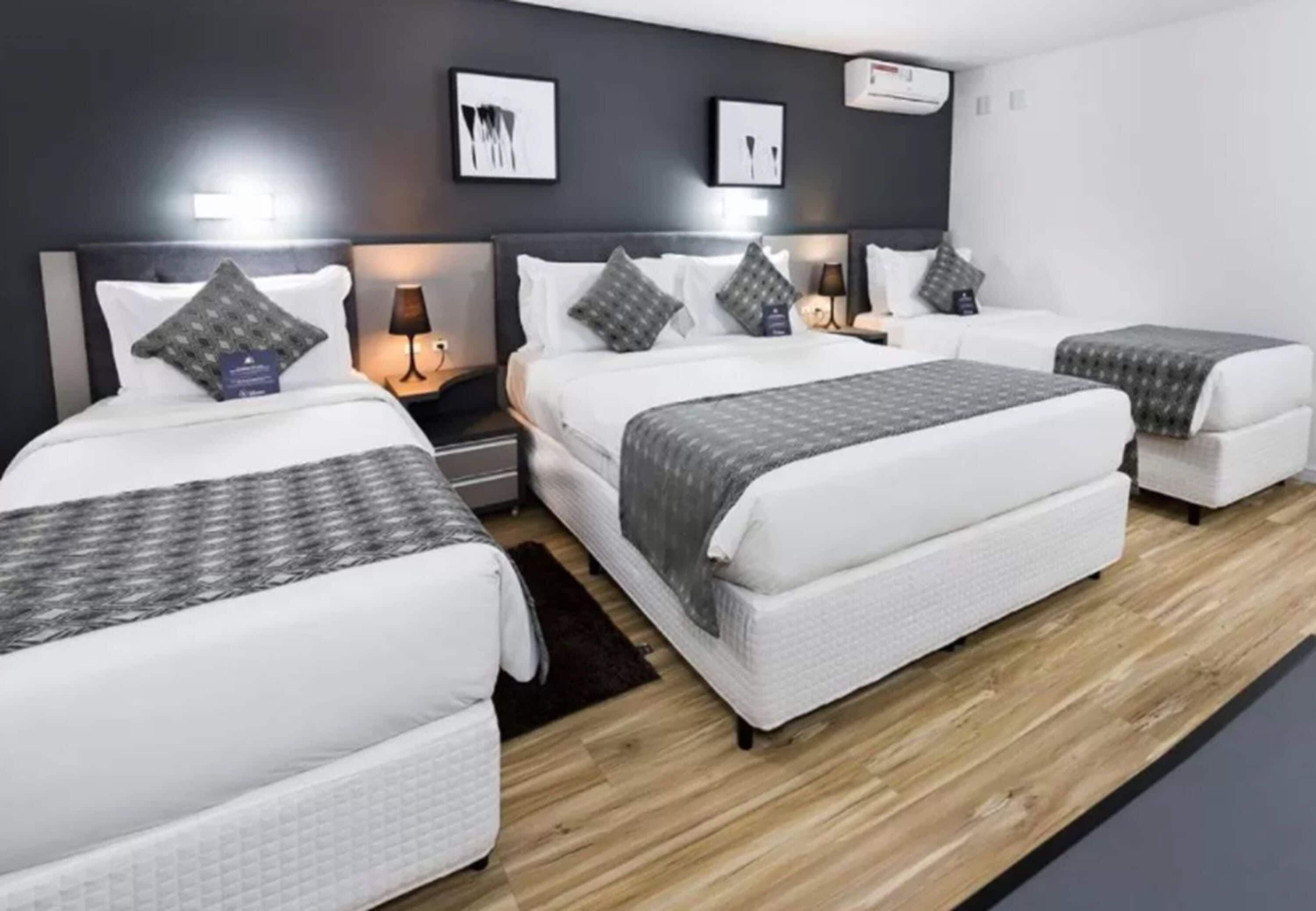 Ein Hotelzimmer mit drei nebeneinander stehenden Betten, jeweils mit weißer Bettwäsche und grauen Überwürfen. An den Nachttischen stehen Lampen und es ist eine Wand-Klimaanlage angebracht.