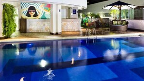 Una piscina con solárium de azulejos, una recepción con un mural colorido y una zona de estar con una silla de mimbre y paraguas.