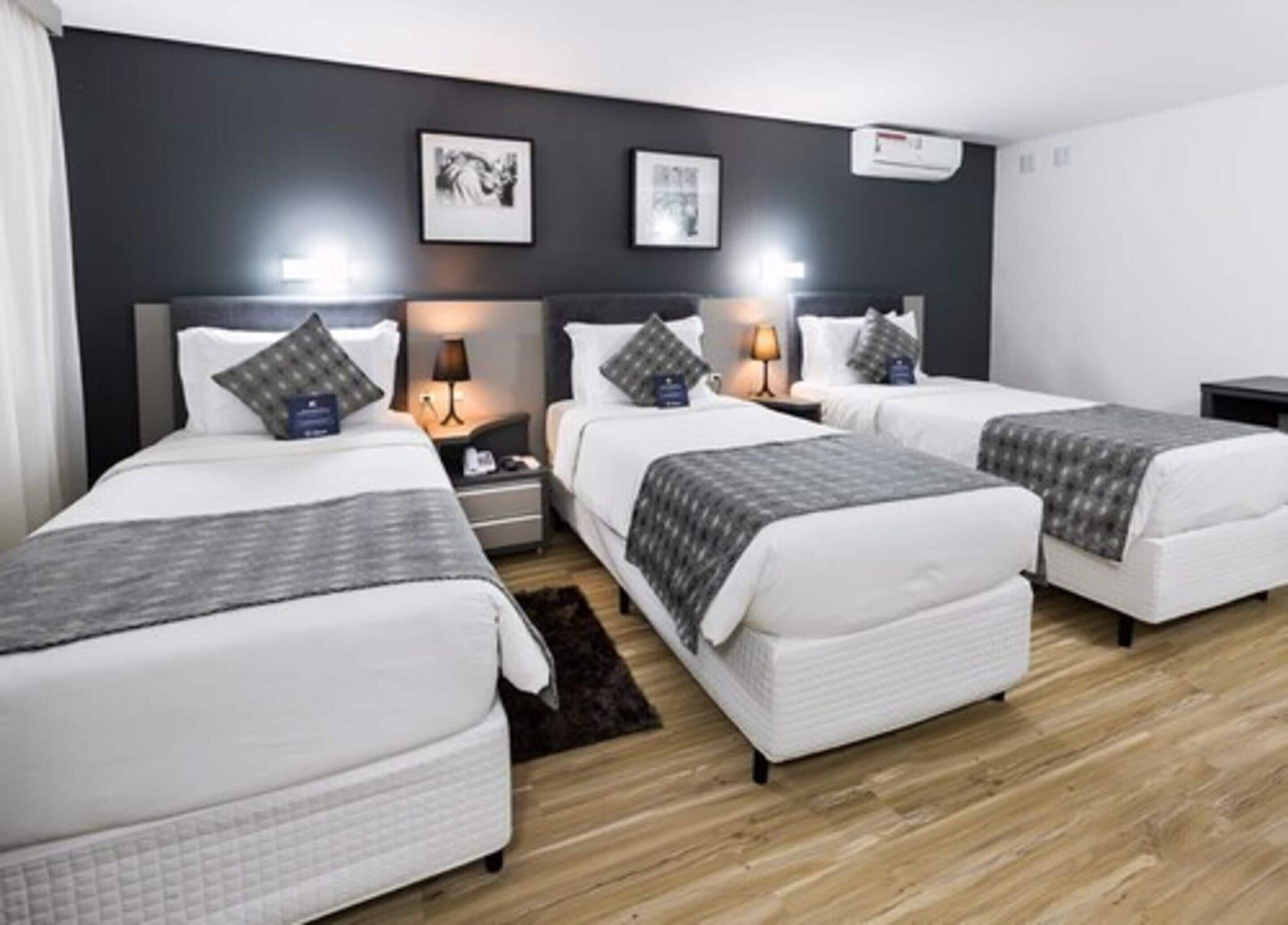 A hotel room with four single beds, a dark wall with two framed pictures, a small rug, and a wooden floor.
